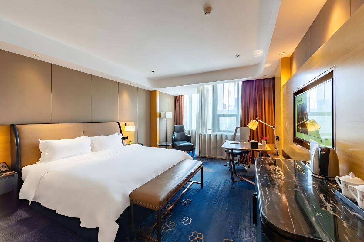 A hotel room with a large bed, a desk, a chair, a bench, and a TV.