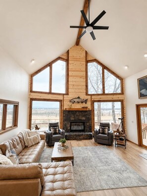 A spacious living room with a stone fireplace, leather sofas, and large windows offering views of the outdoors.