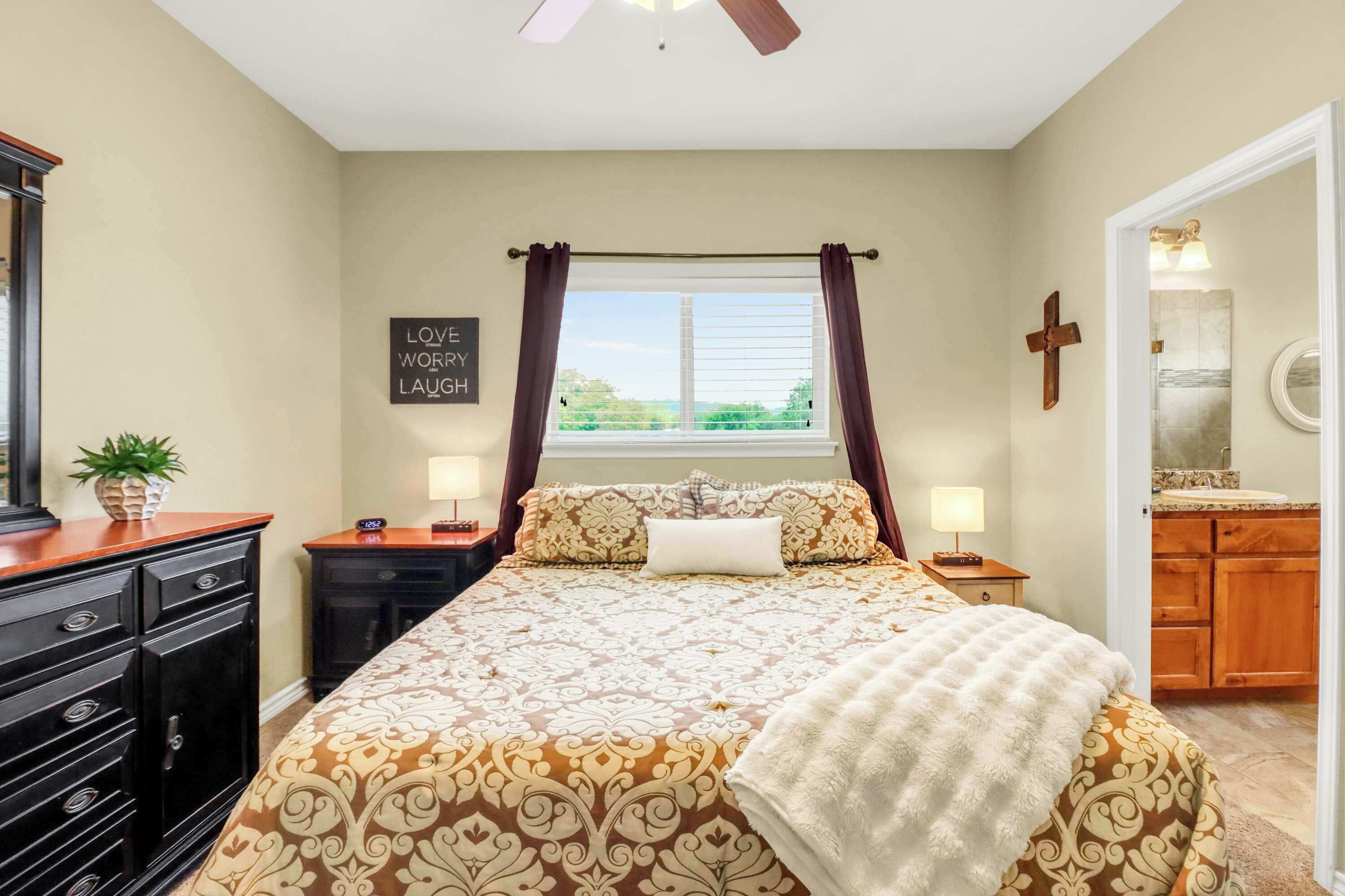 A bedroom with a bed, nightstands, a dresser, a window with curtains, a wall with a cross, and a bathroom visible through an open door.