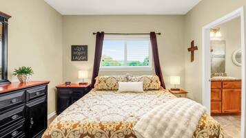 A bedroom with a bed, nightstands, a dresser, a window with curtains, a wall with a cross, and a bathroom visible through an open door.