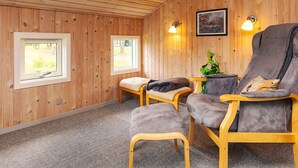 Une pièce aux murs en bois, avec deux fenêtres, un canapé, un fauteuil et une petite table sur laquelle se trouve une plante.