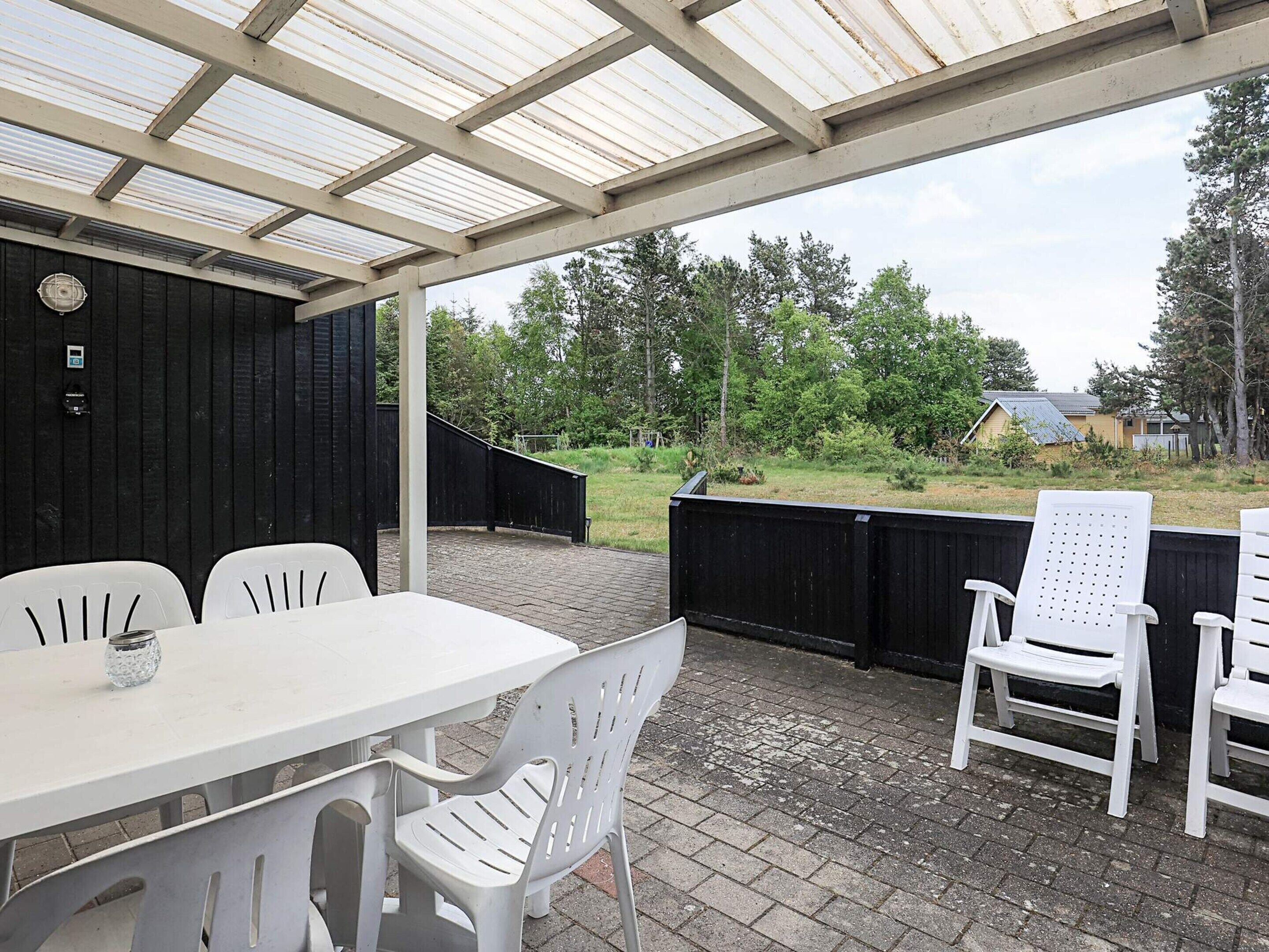 Un patio al aire libre con una mesa y sillas, una zona cubierta con techo metálico y vistas a un área boscosa.