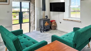 Une pièce avec une cheminée, des fauteuils à revêtement vert, une table en bois, un téléviseur fixé au mur et une vue à travers une porte vitrée.
