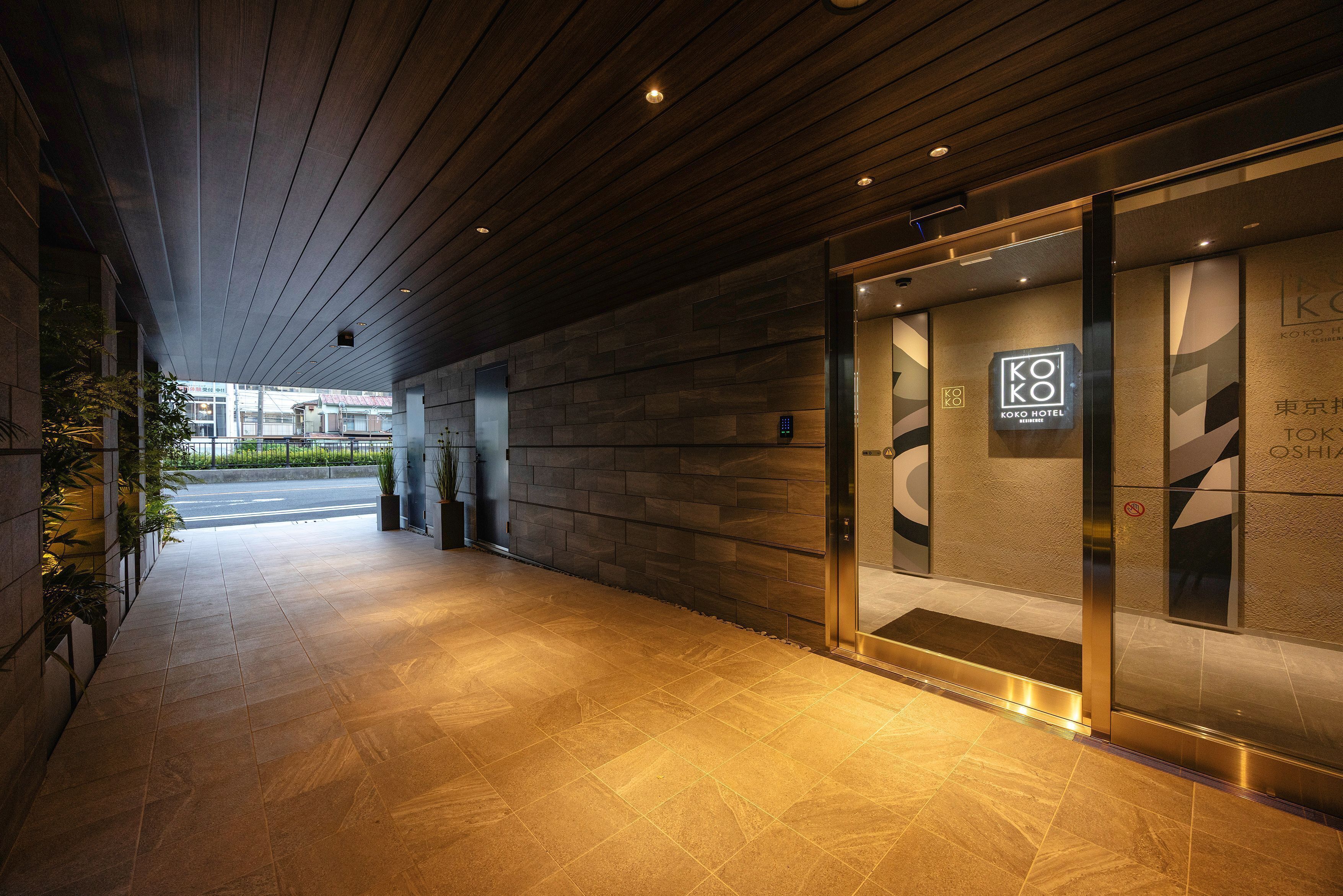 A hotel entrance with a modern design, featuring a wooden ceiling, a glass door with the hotel's logo, and a tiled floor.