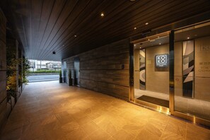 A hotel entrance with a modern design, featuring a wooden ceiling, a glass door with the hotel's logo, and a tiled floor.
