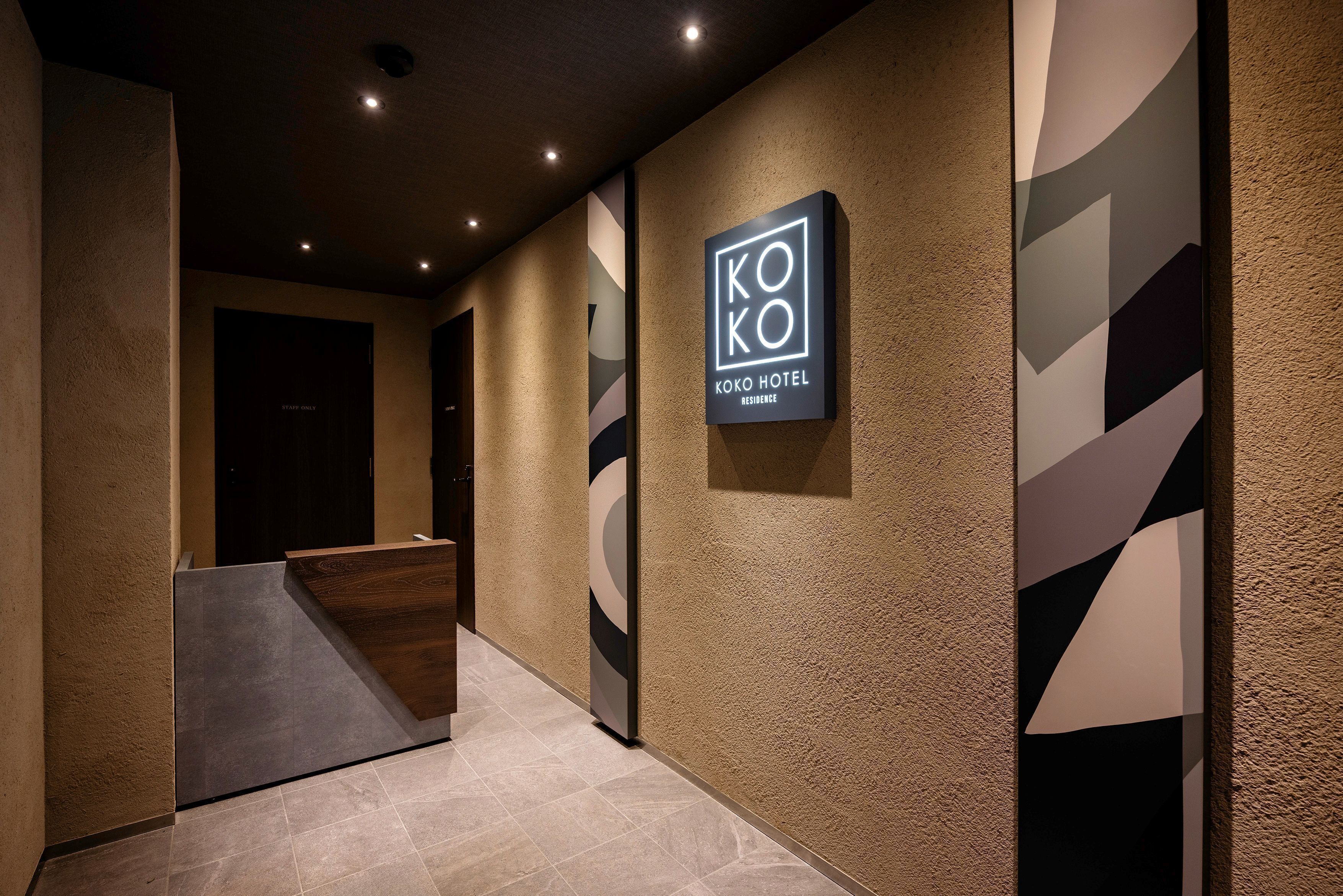 Ein Hotel-Empfangsbereich mit modernem Design, der einen Empfangstresen, ein Schild mit der Aufschrift 'KOKO HOTEL RESIDENCE' und eine strukturierte Wand zeigt.