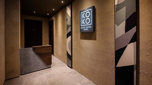 Zona de recepción de hotel con diseño moderno, que incluye un mostrador de recepción, un cartel con la leyenda 'KOKO HOTEL RESIDENCE' y una pared con textura.