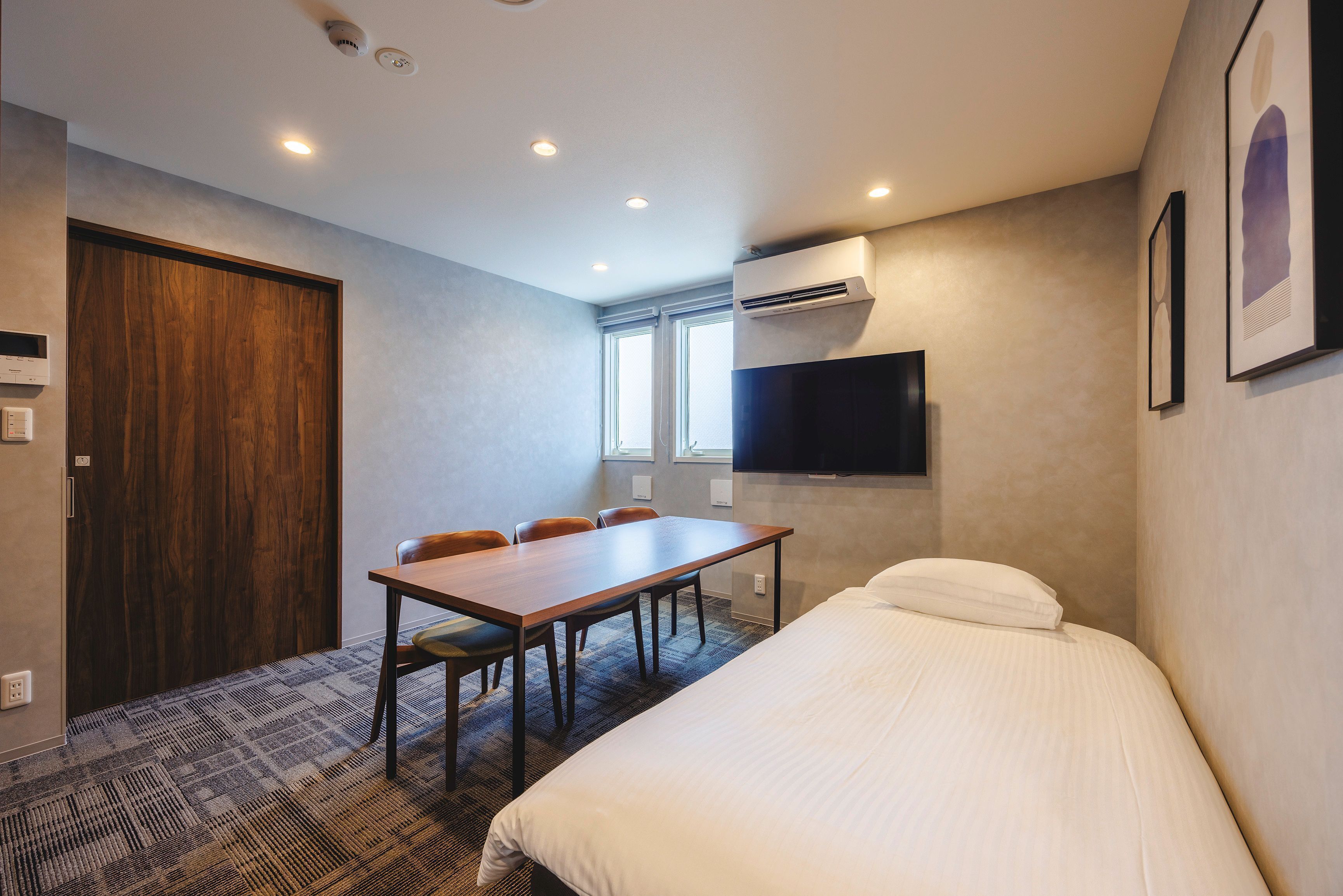 A hotel room with a bed, a desk, and a chair. There is a window, an air conditioning unit, and a television mounted on the wall.