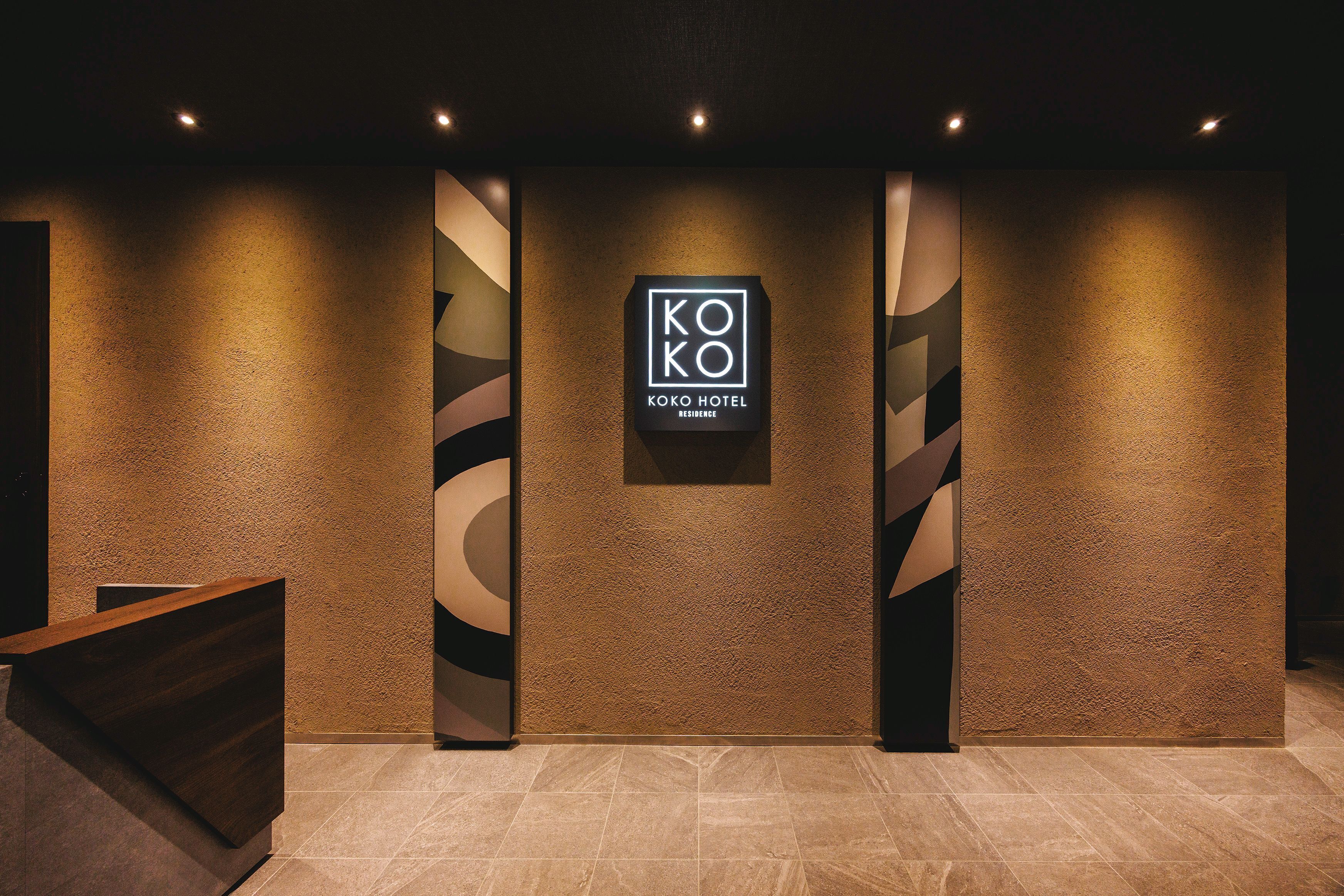 Un espace d’accueil d’hôtel au design moderne, comprenant un comptoir d’accueil, une enseigne indiquant « KOKO HOTEL » et un mur à texture.