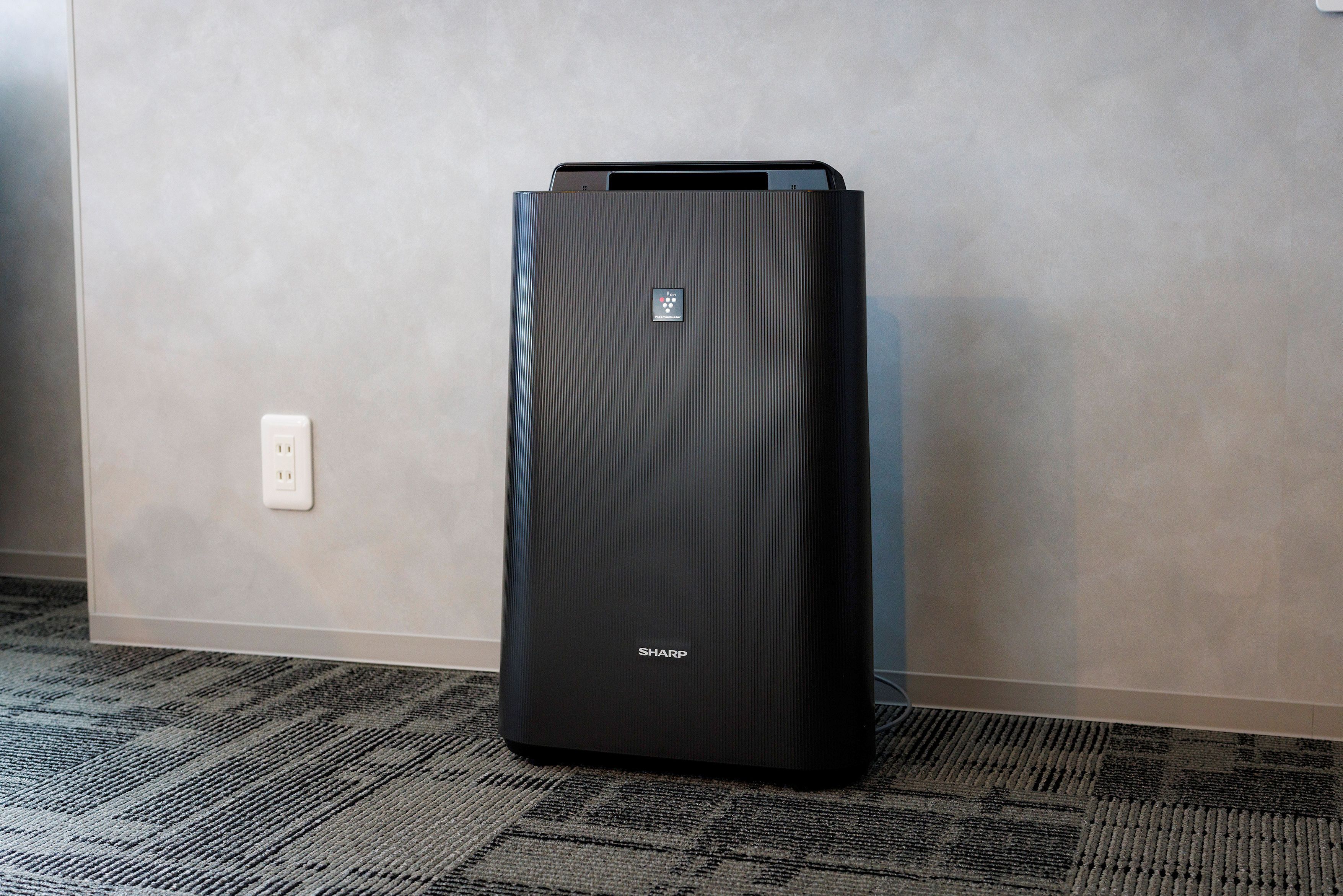 A black, rectangular, metallic object with a label on the front, standing on a carpeted floor against a wall with a power outlet.