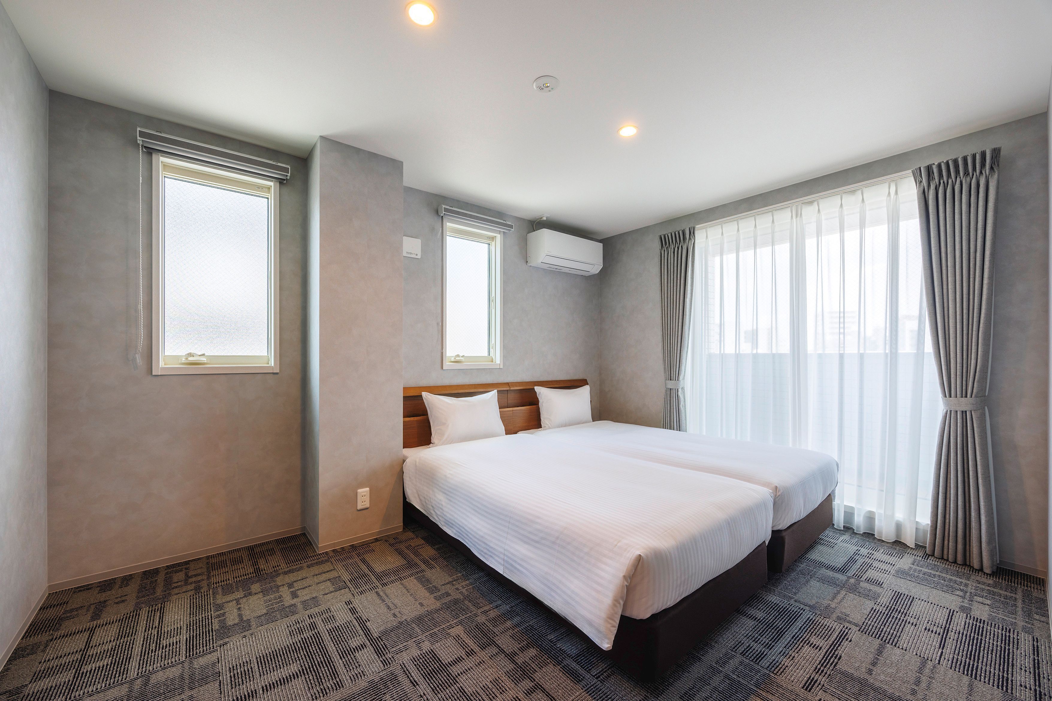 Habitación de hotel con cama doble, dos ventanas, aire acondicionado y piso alfombrado.