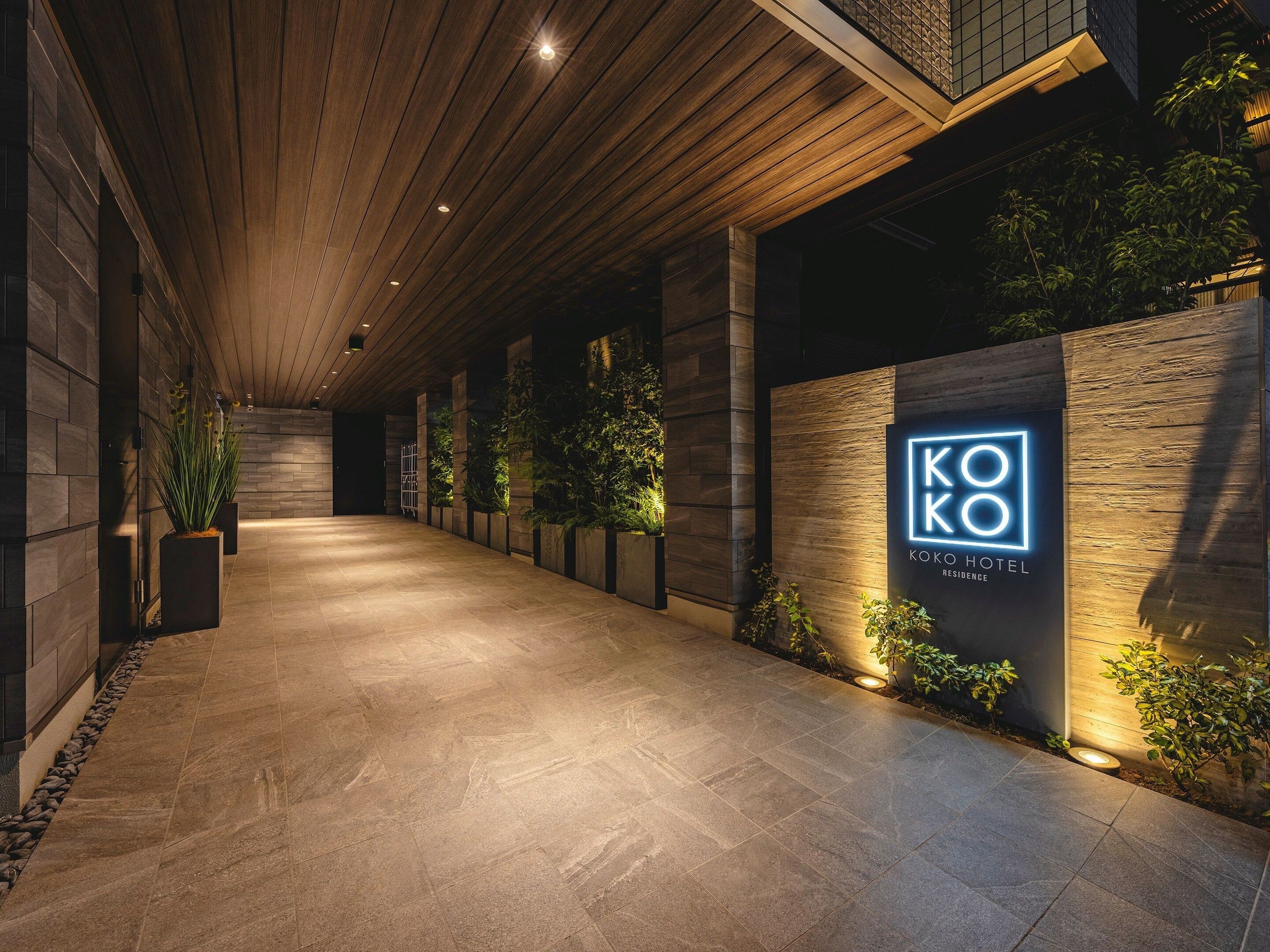 Een gang met een houten plafond, een bord met 'KOKO HOTEL RESIDENCE' en potplanten langs de muren.