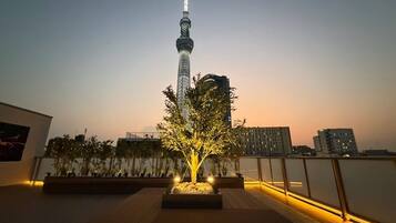 Une vue nocturne d’une ville, avec une tour imposante, un espace paysager agrémenté d’un arbre et un bâtiment doté d’un balcon.