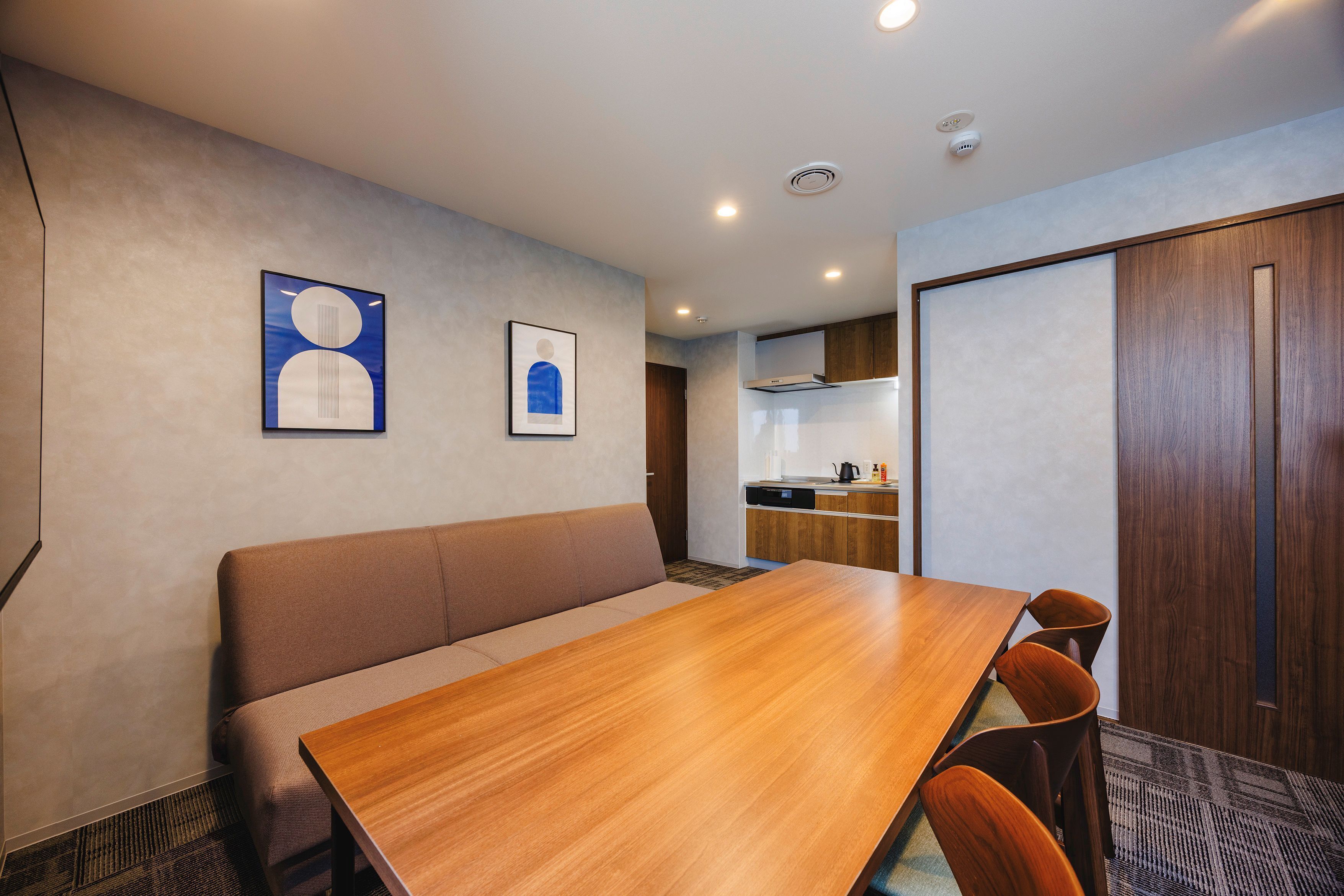 A modern hotel room with a wooden dining table, a sofa, and a kitchenette in the background.