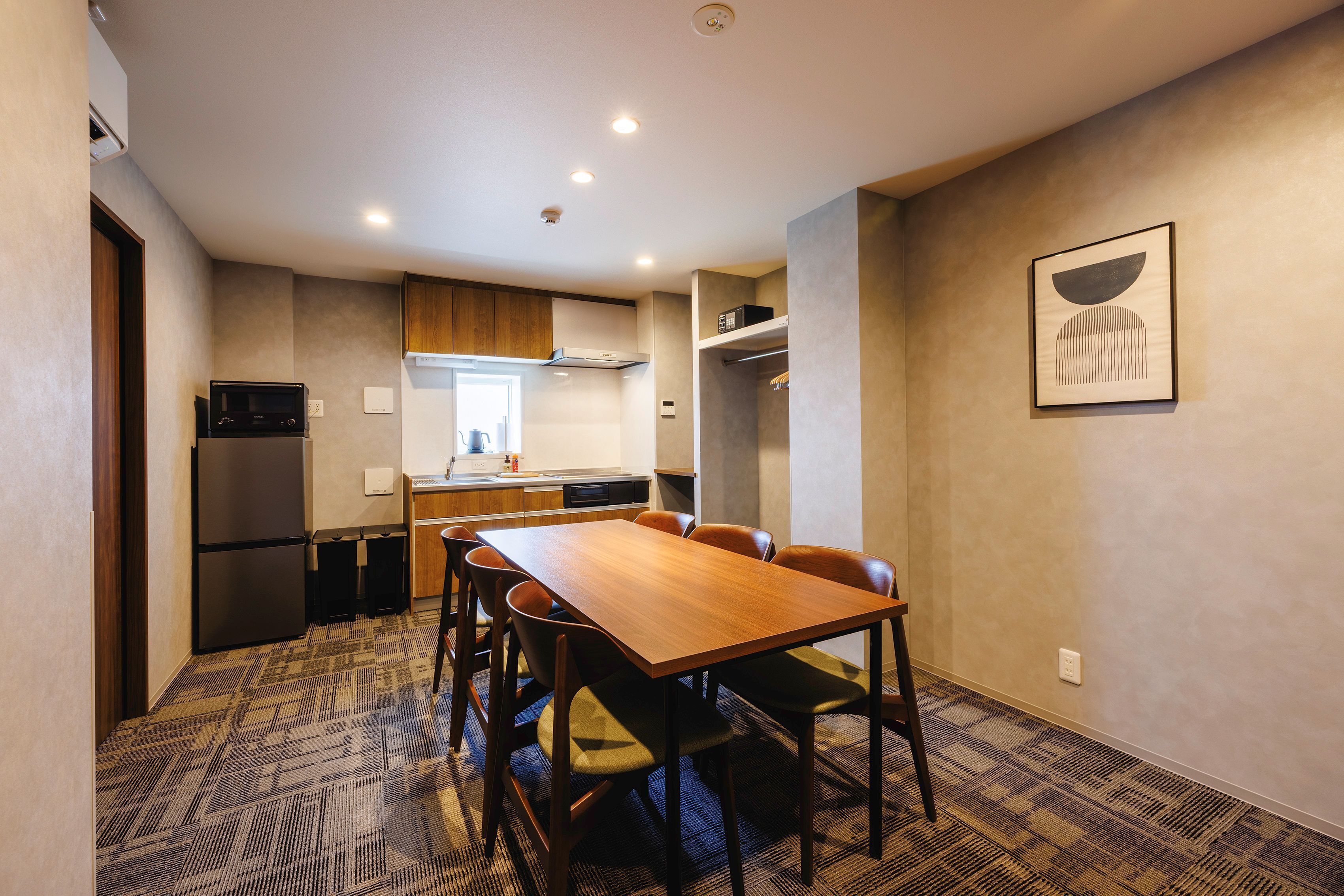 Een compacte hotelkamer met een kitchenette, een eethoek met een houten tafel en stoelen, en een enkel kunstwerk aan de muur.