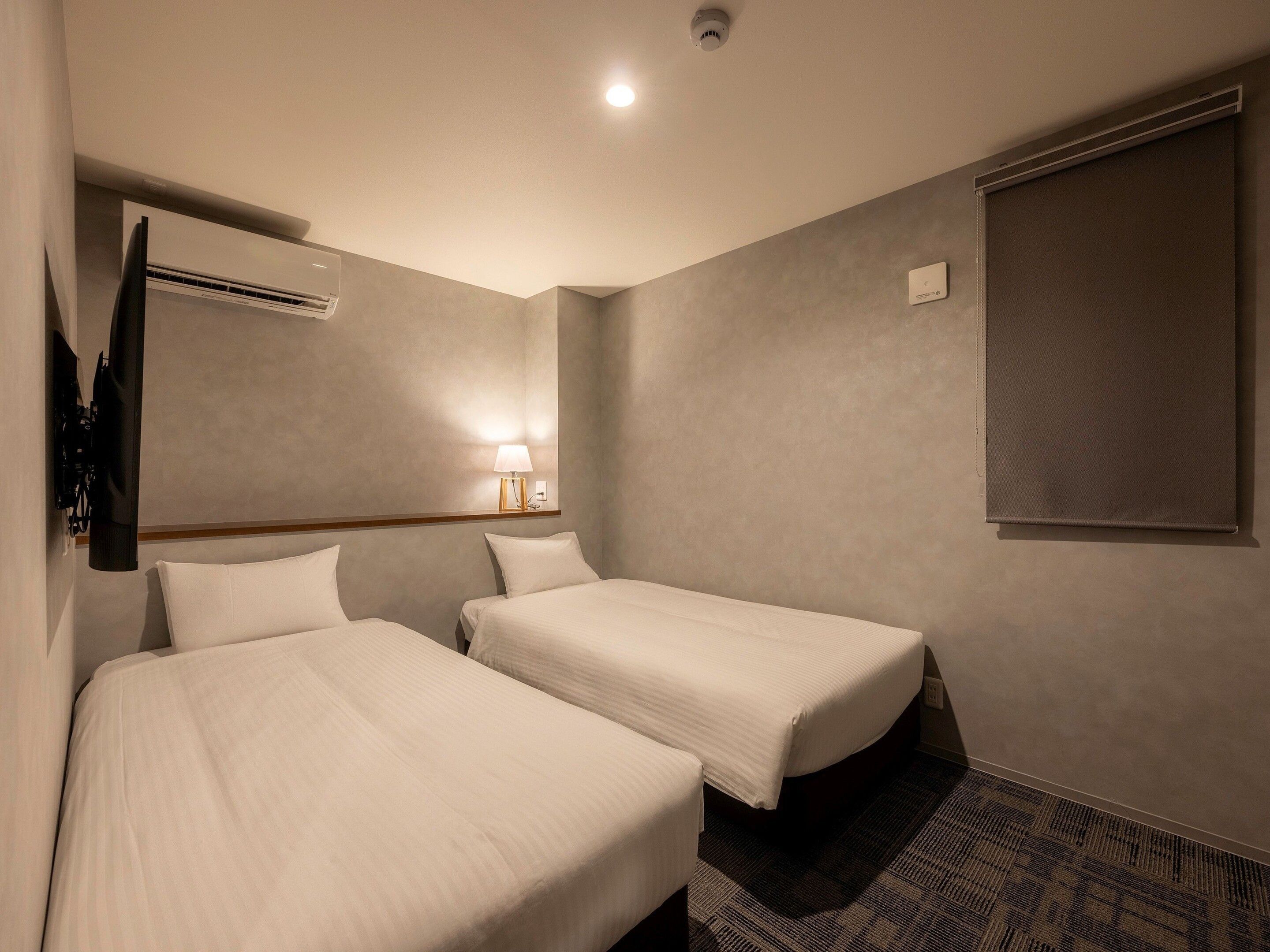 Habitación de hotel con dos camas, un televisor de pantalla plana montado en la pared, un equipo de aire acondicionado, una lámpara de noche y una ventana con persiana.