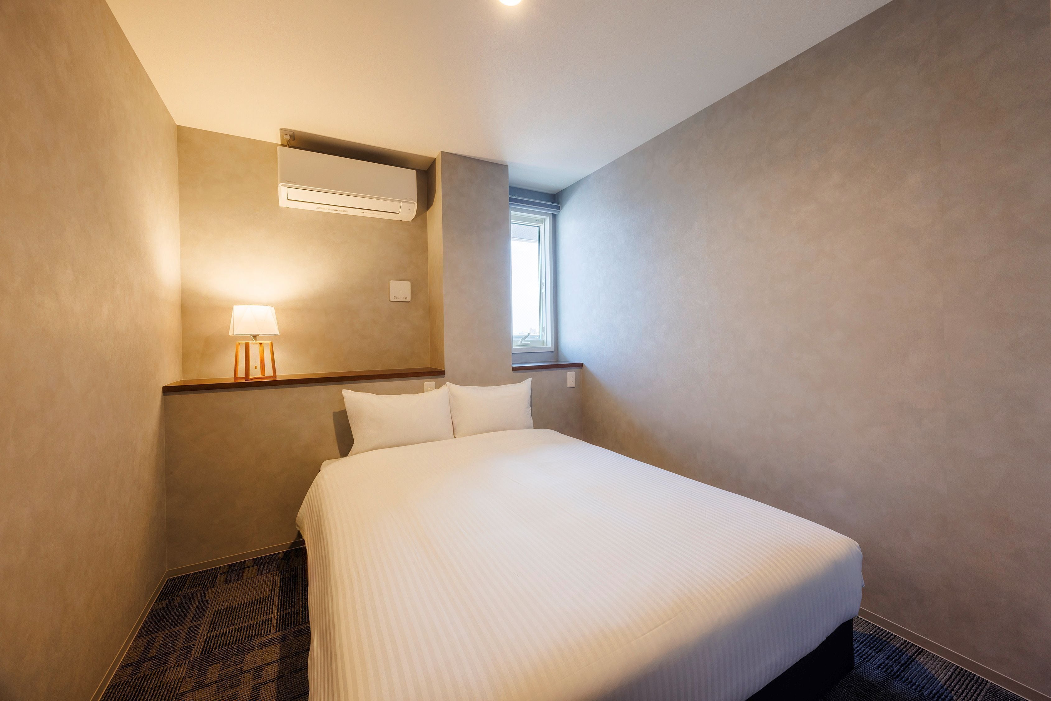 Une chambre d’hôtel avec un lit, un climatiseur fixé au mur, une lampe de chevet et une fenêtre.
