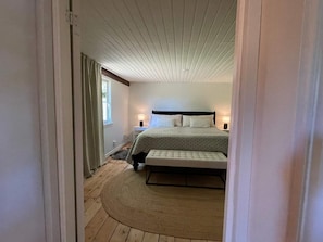 Ett sovrum med en säng, sängbord, en bänk, ett fönster och ett tak med synliga träbjälkar.