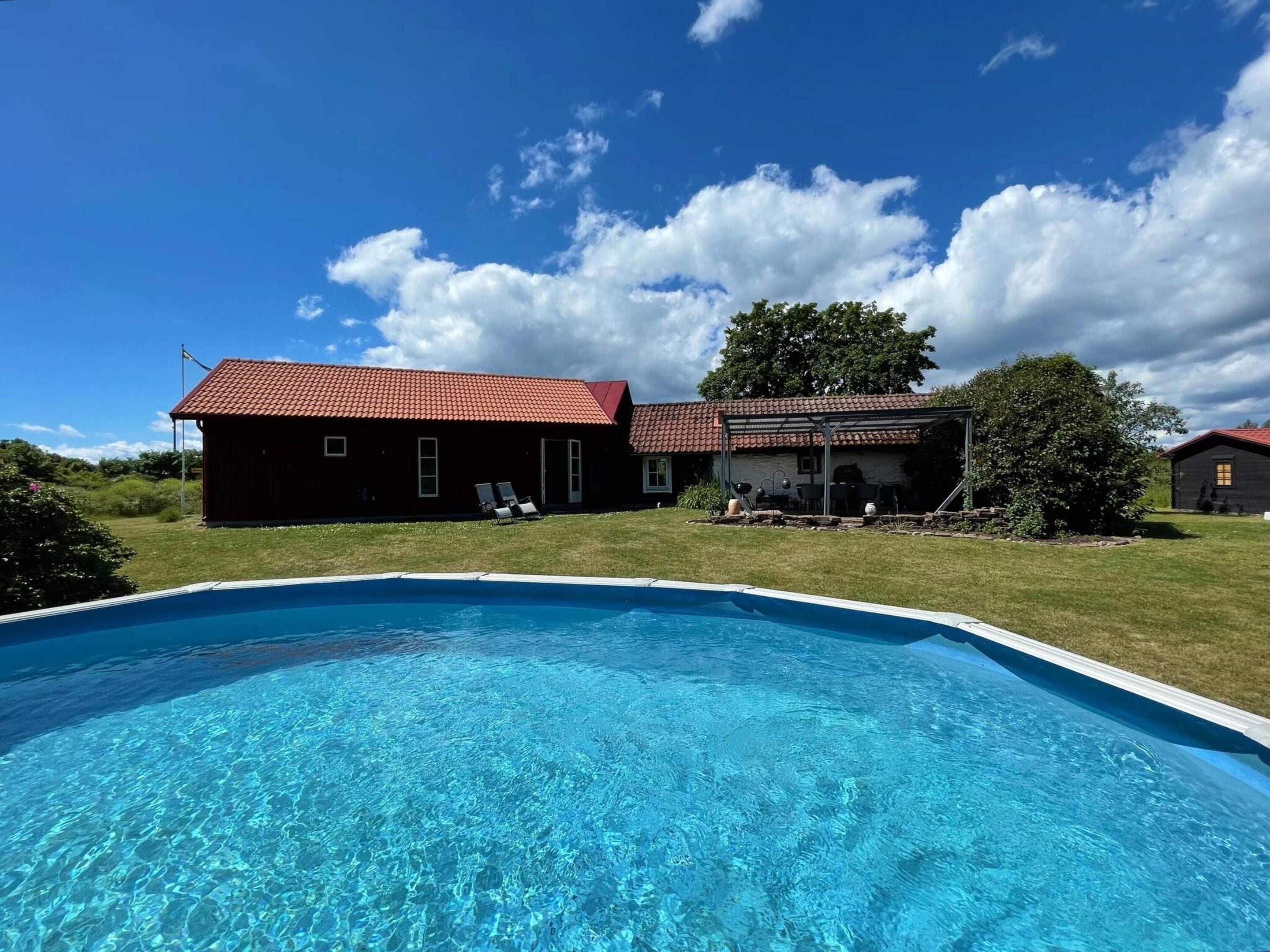 Une piscine à l’eau d’un bleu limpide, entourée d’une pelouse, et deux bâtiments aux toits couverts de tuiles rouges en arrière-plan.