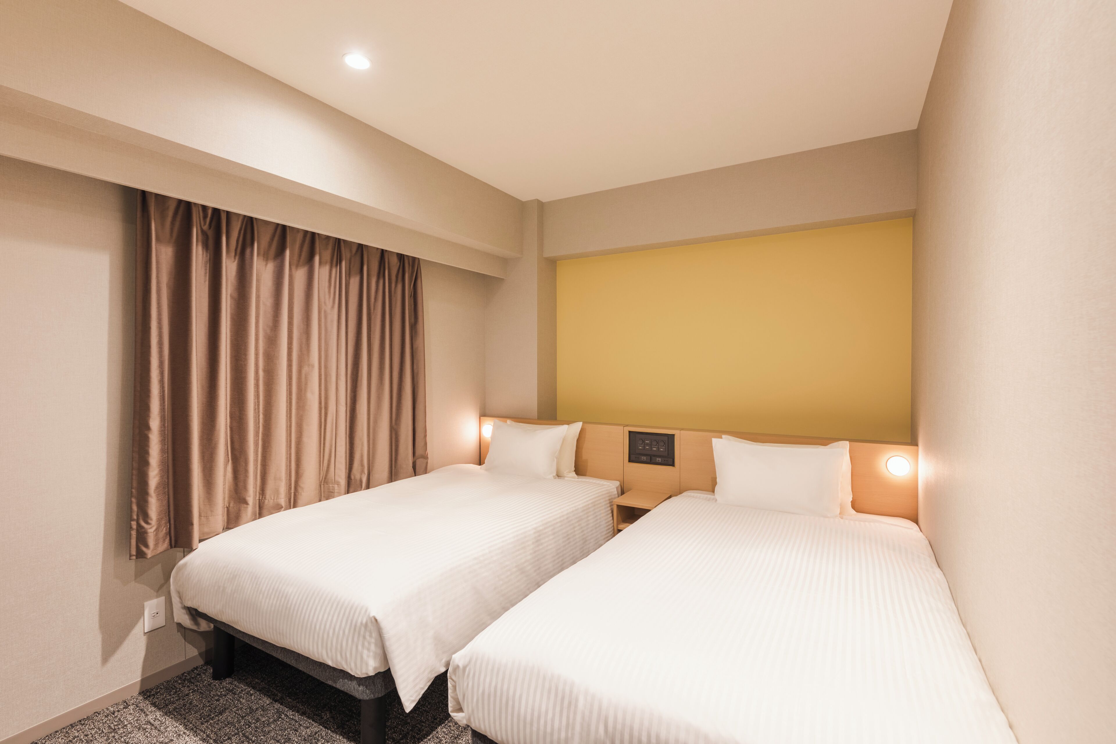 A hotel room with two beds, a headboard with a control panel, a window with curtains, and a wall-mounted light.