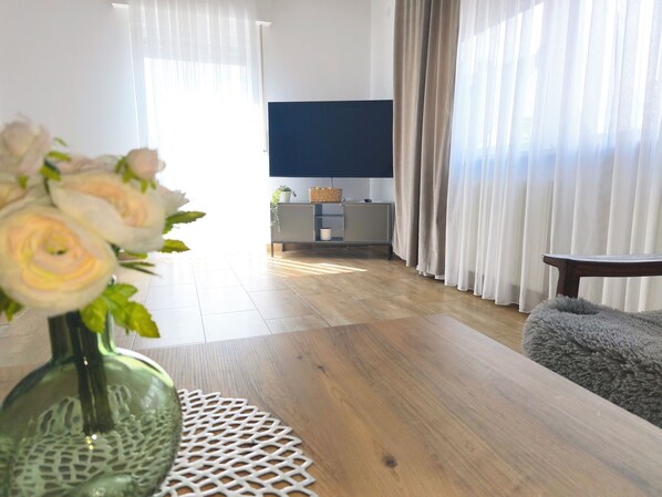 A living room with a wooden floor, a TV on the wall, a grey sofa, and a vase with flowers on a table.