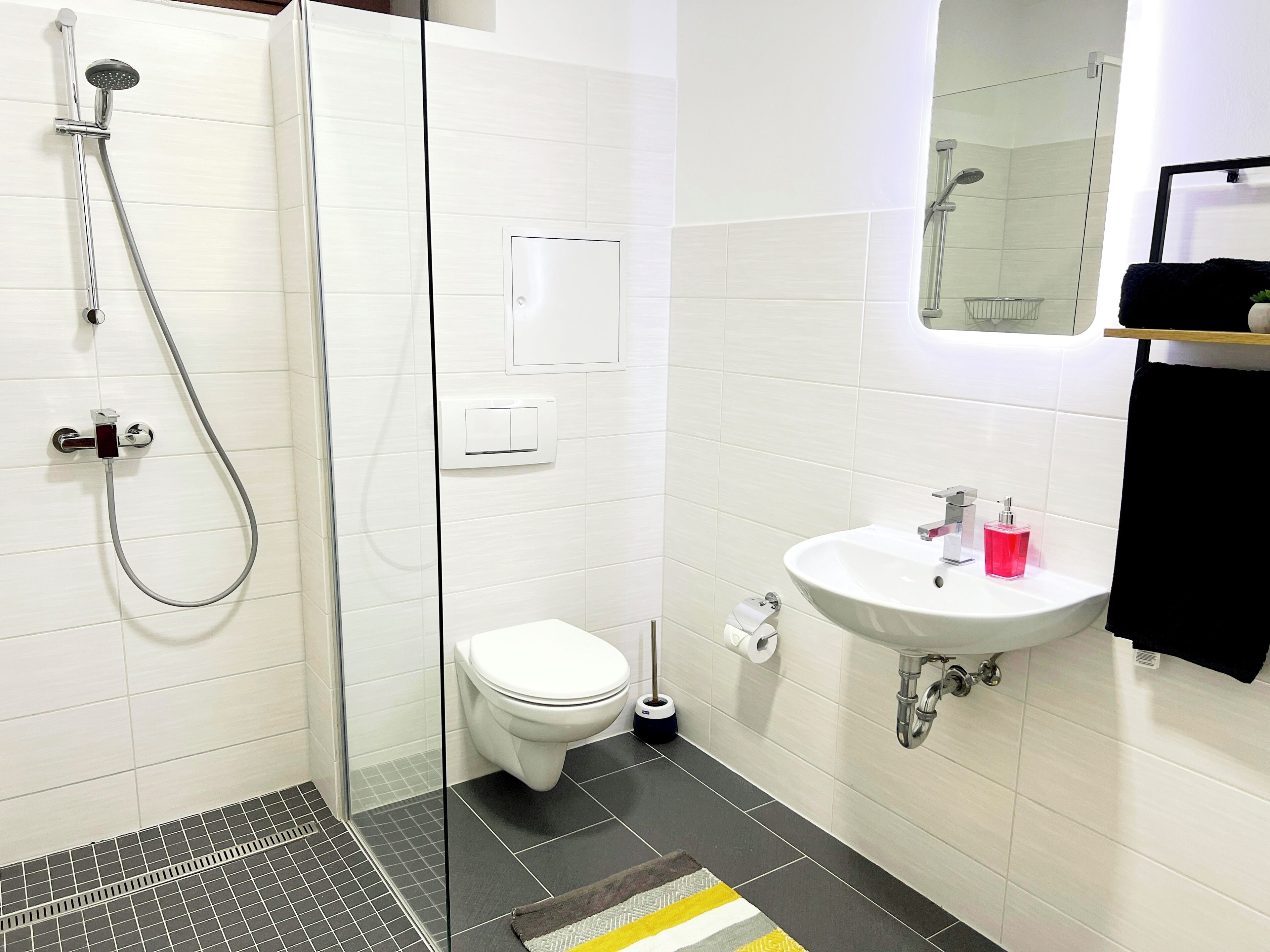 Ein modernes Badezimmer mit einer Duschkabine aus Glas, einer Toilette, einem Waschbecken mit Spiegel und einem schwarzen Handtuch an einem Ablagegestell.