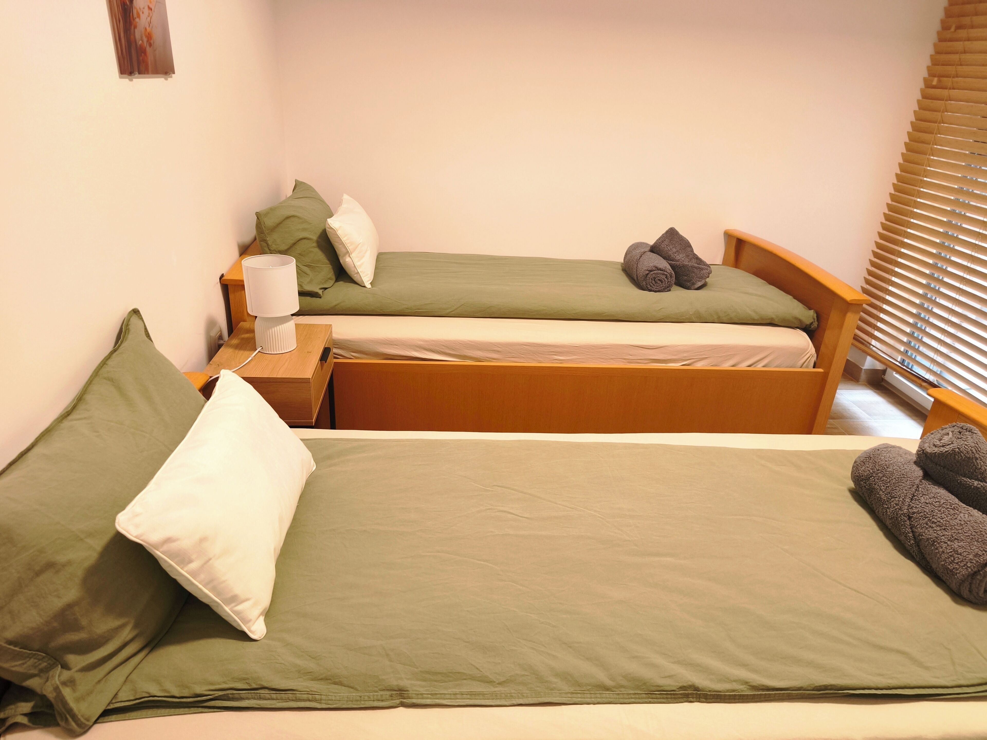 Two single beds with green bedding, a bedside table with a lamp, and a window with blinds.