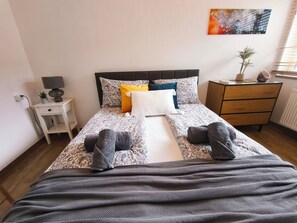 Ein ordentlich bezogenes Bett mit gemusterten Betttextilien, ein Nachttisch mit Lampe und eine Kommode mit einer Pflanze.