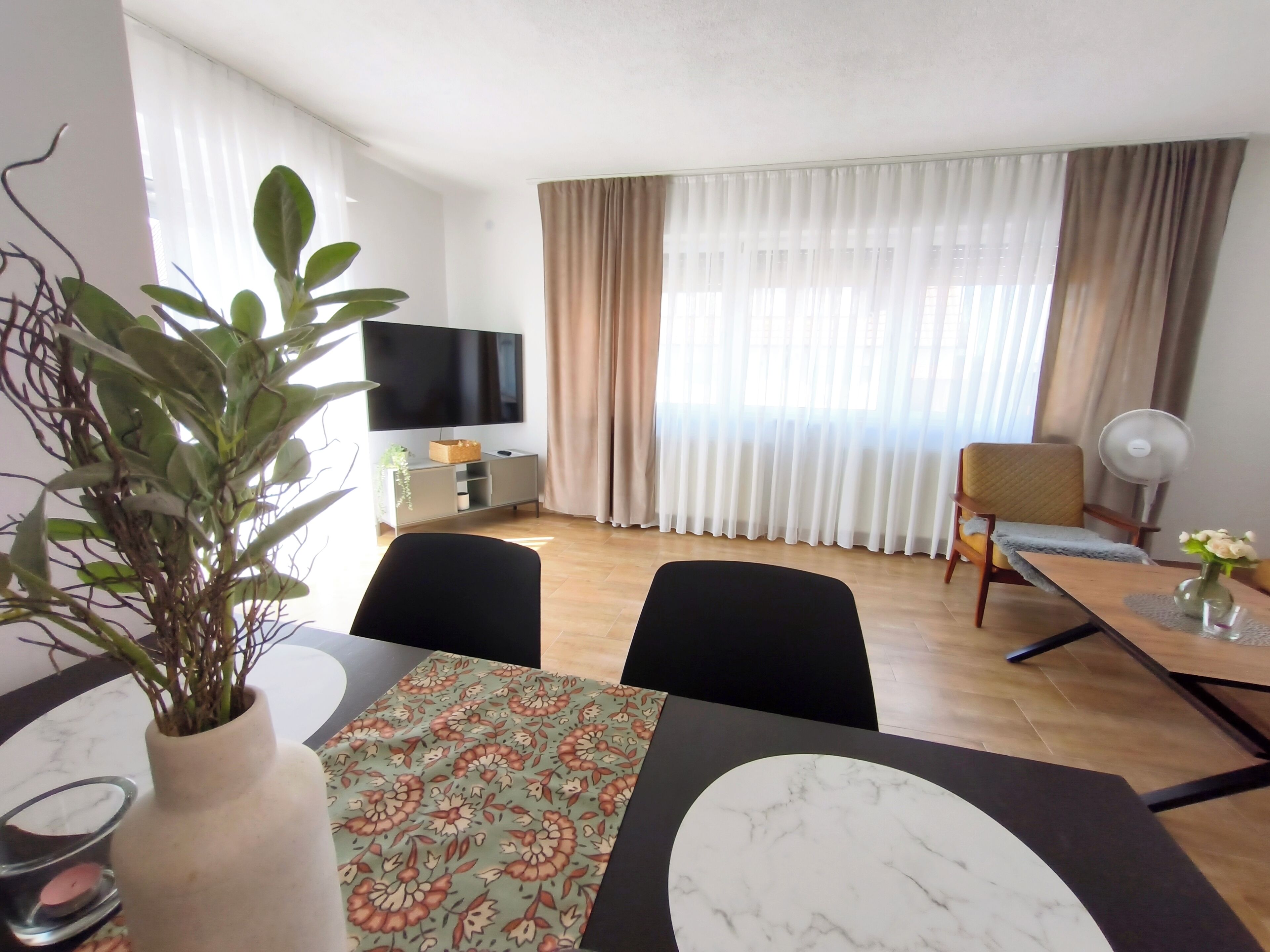 Ein modernes Hotelzimmer mit Essbereich, einem Flachbildfernseher, einem großen Fenster mit Vorhängen, einem Ventilator und einer Zimmerpflanze.