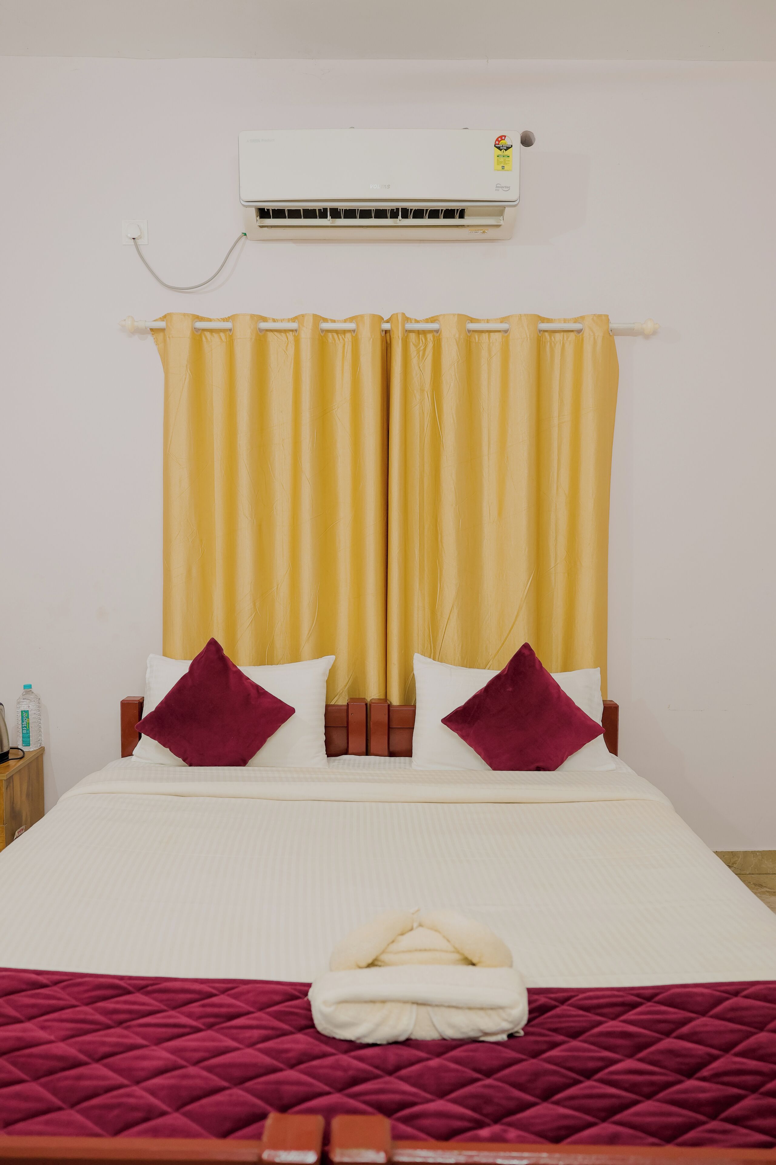 Ein Hotelzimmer mit Bett, Vorhängen, Kissen, einer Decke, einer Klimaanlage und einem Nachttisch mit einer Flasche.