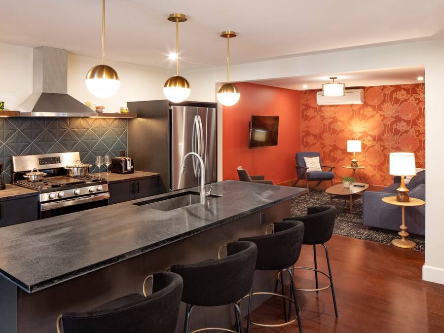 A modern kitchen with a central island, bar stools, a refrigerator, and a dining area with a sofa and a table.