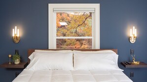 A bedroom with a bed, two bedside tables, a window with a view of autumn trees, and wall-mounted sconces. - Shelburne Springs Hotel (Shelburne)