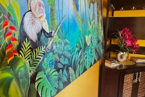 Une pièce ornée d’une grande fresque représentant un singe tenant une épée, avec une plante en pot, et une petite table sur laquelle se trouvent des livres et un vase de fleurs.