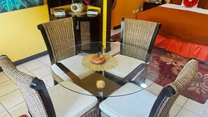 Una mesa con cubierta de vidrio, cuatro sillas de mimbre, un adorno pequeño sobre la mesa y una cama con un cojín rojo al fondo.