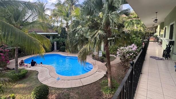 Piscina rodeada de palmeras y follaje, con un paseo cubierto y zona de estar al aire libre.