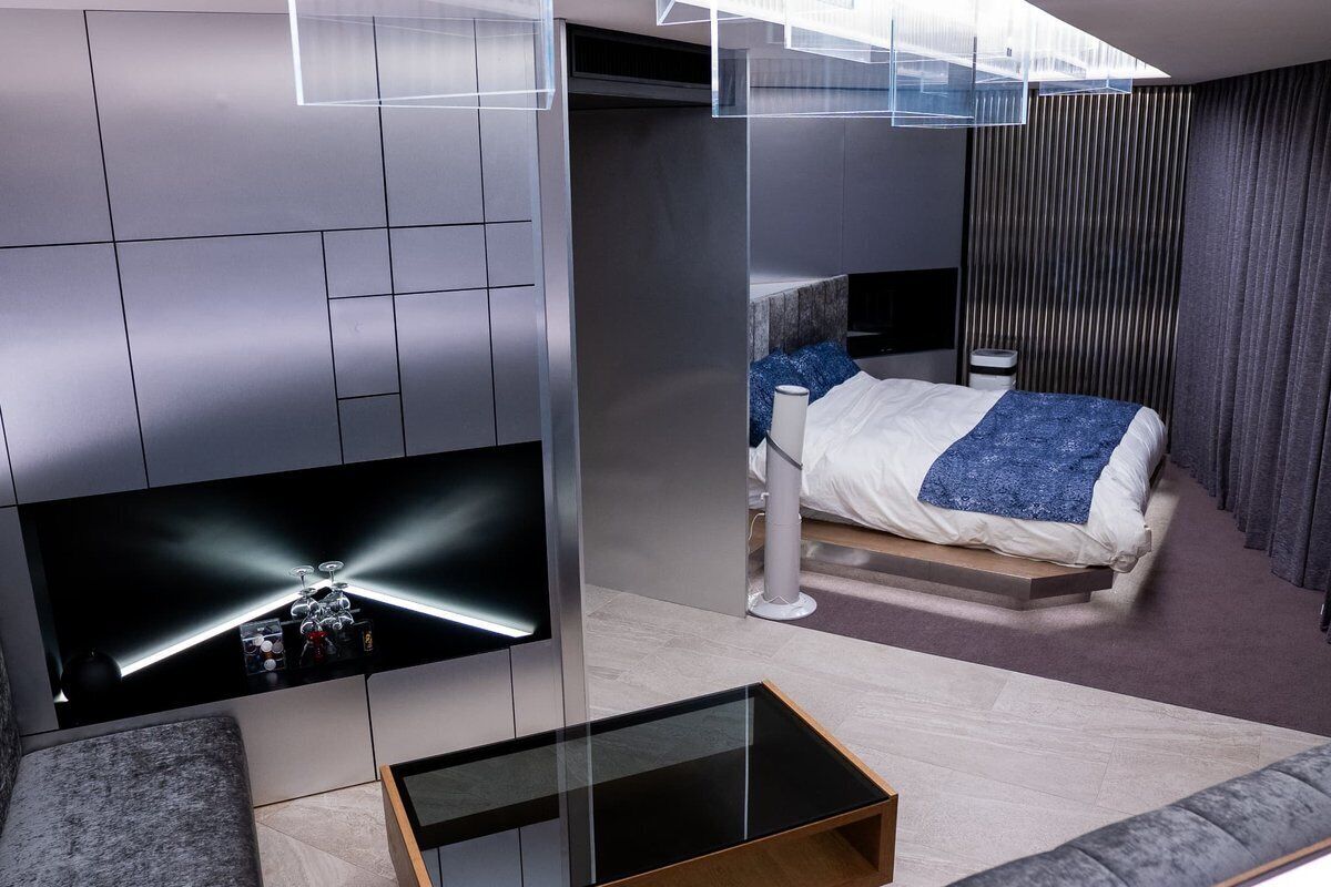 A modern hotel room with a bed, a TV mounted on the wall, a glass coffee table, and a sofa.