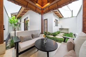A covered outdoor seating area with a sofa and a round table, surrounded by greenery and a small garden.