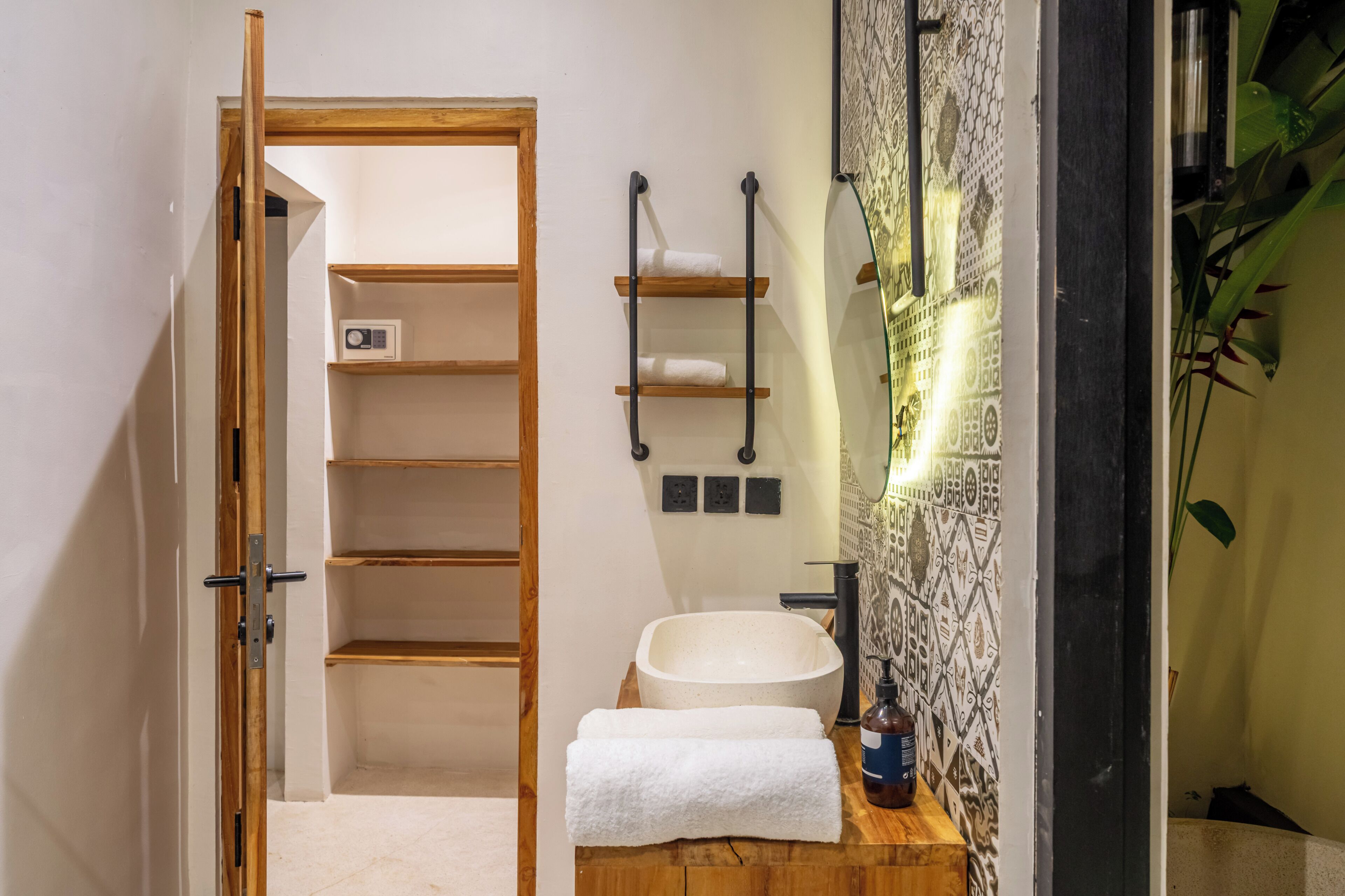 A bathroom with a wooden door, a white sink, a towel, a shelf with towels, and a patterned wall.