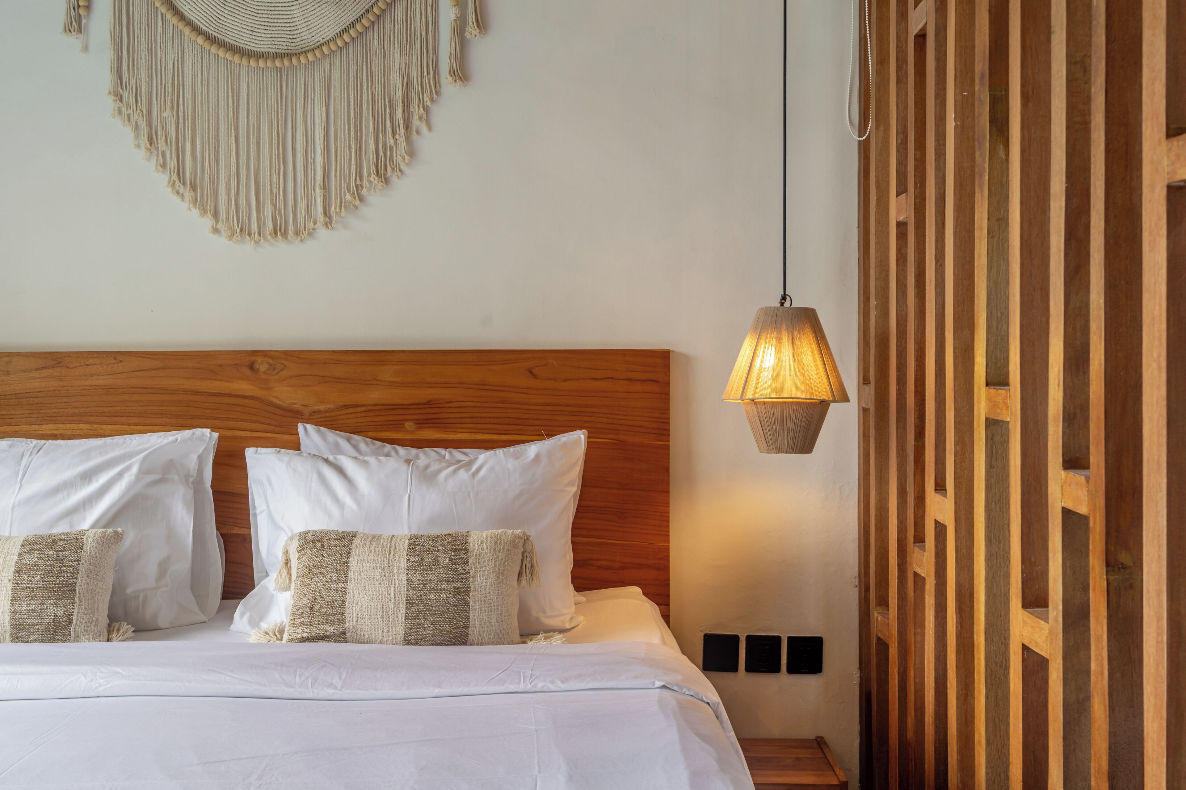 A neatly made bed with white bedding and two decorative pillows, a wooden headboard, a wall-mounted macrame piece, and a hanging lamp.