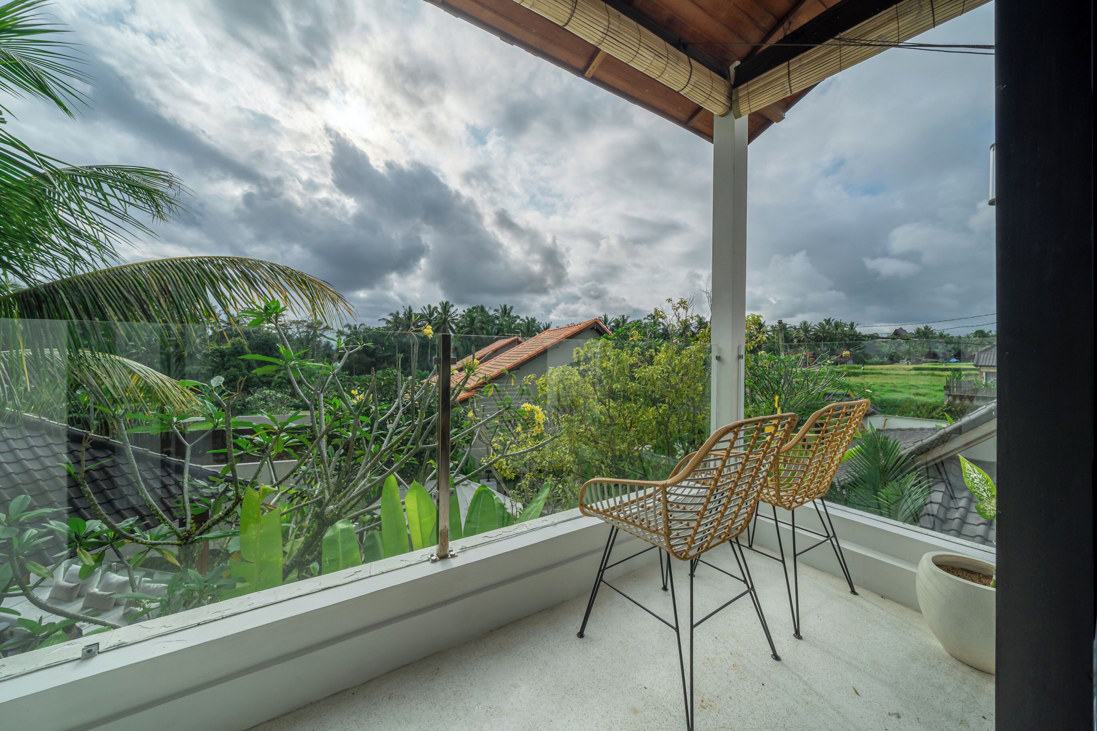 A balcony with two wicker chairs, a potted plant, and a view of a residential area with greenery and a tiled roof.