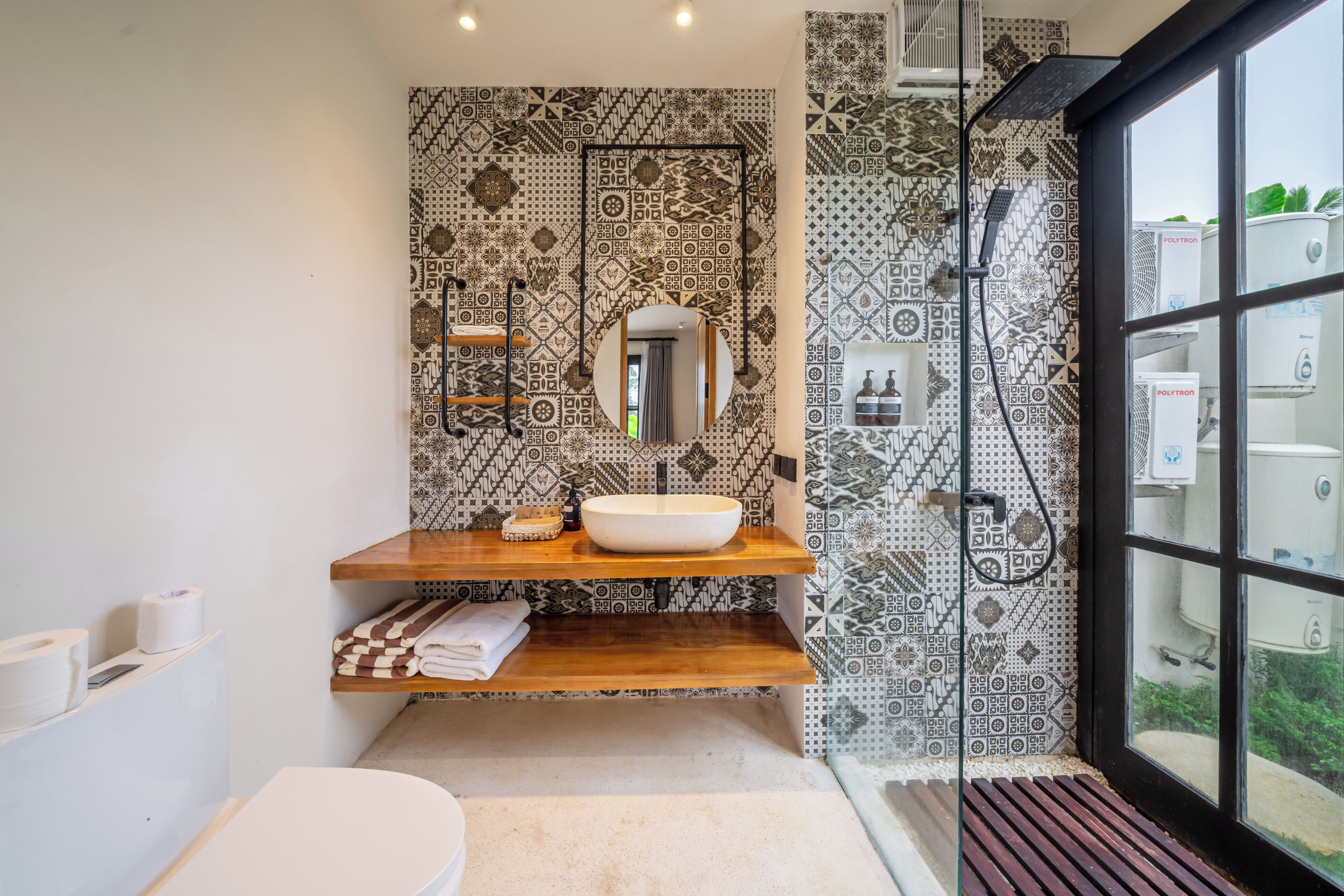 A bathroom with a wooden vanity, a large mirror, patterned tiles, a shower, and a view through a window.