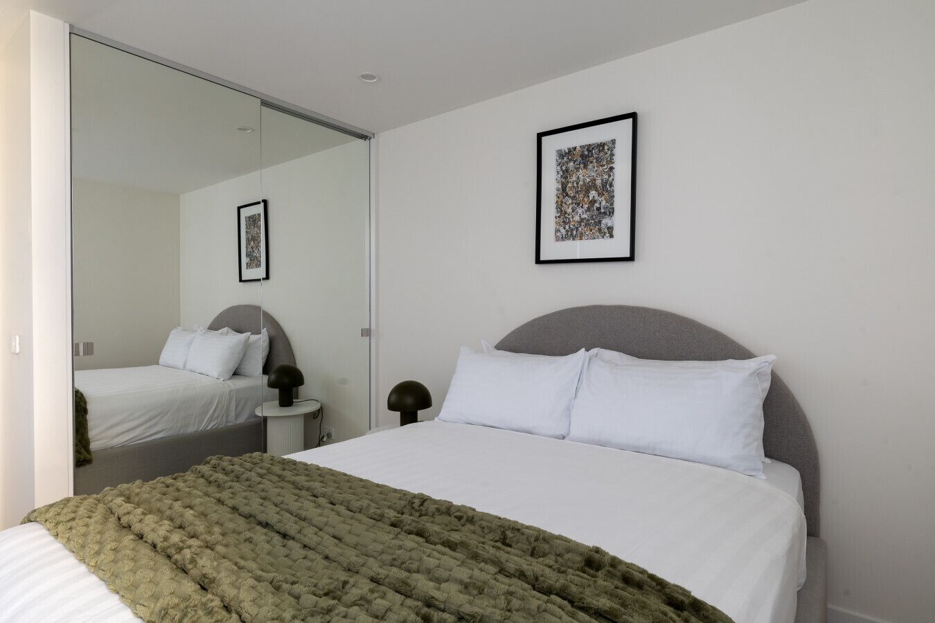 Een slaapkamer met een bed, nachtkastje, lamp, spiegel en een lijst met een afbeelding aan de muur.
