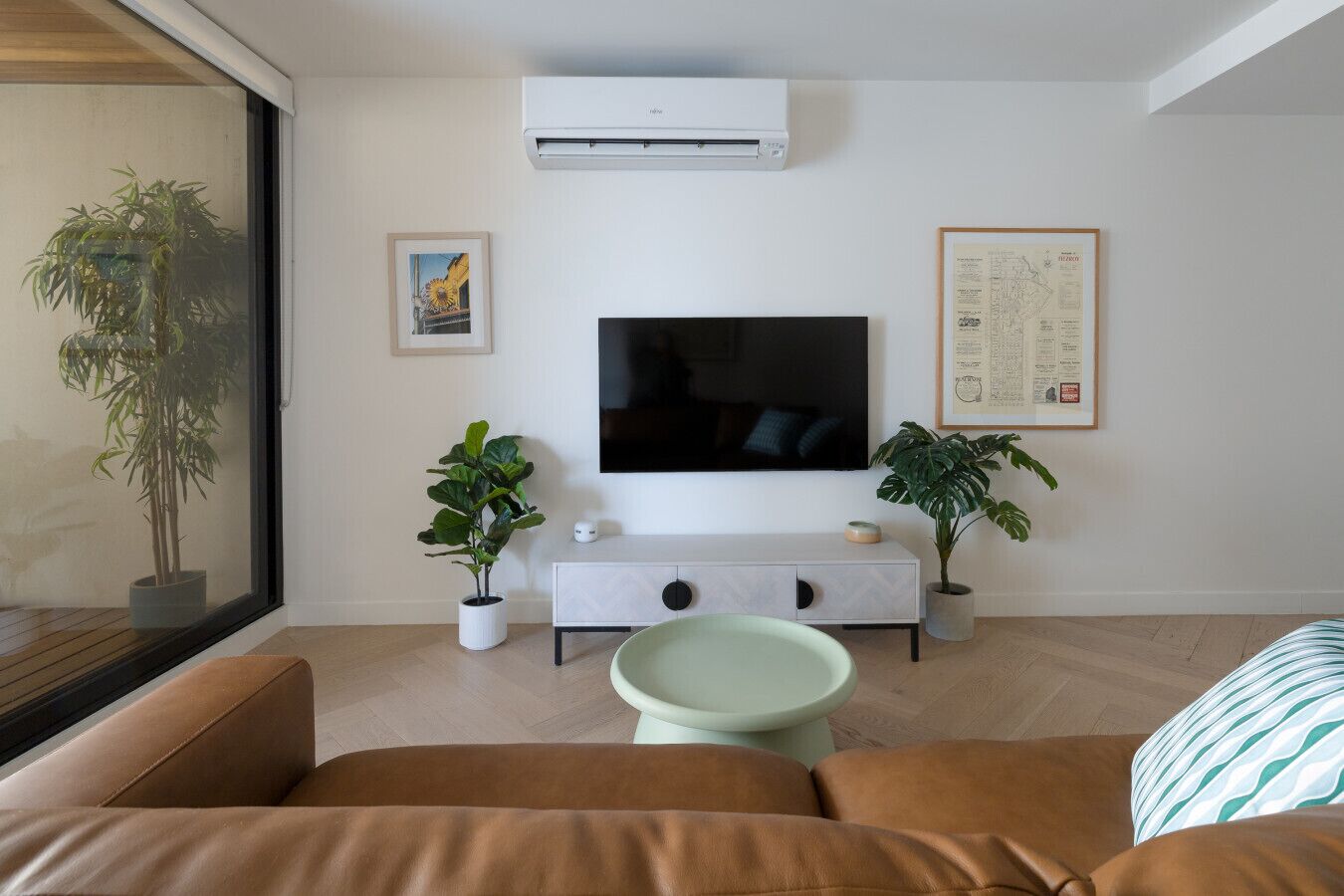 A modern living room with a large flat-screen TV, a white console table, two potted plants, and a large window.
