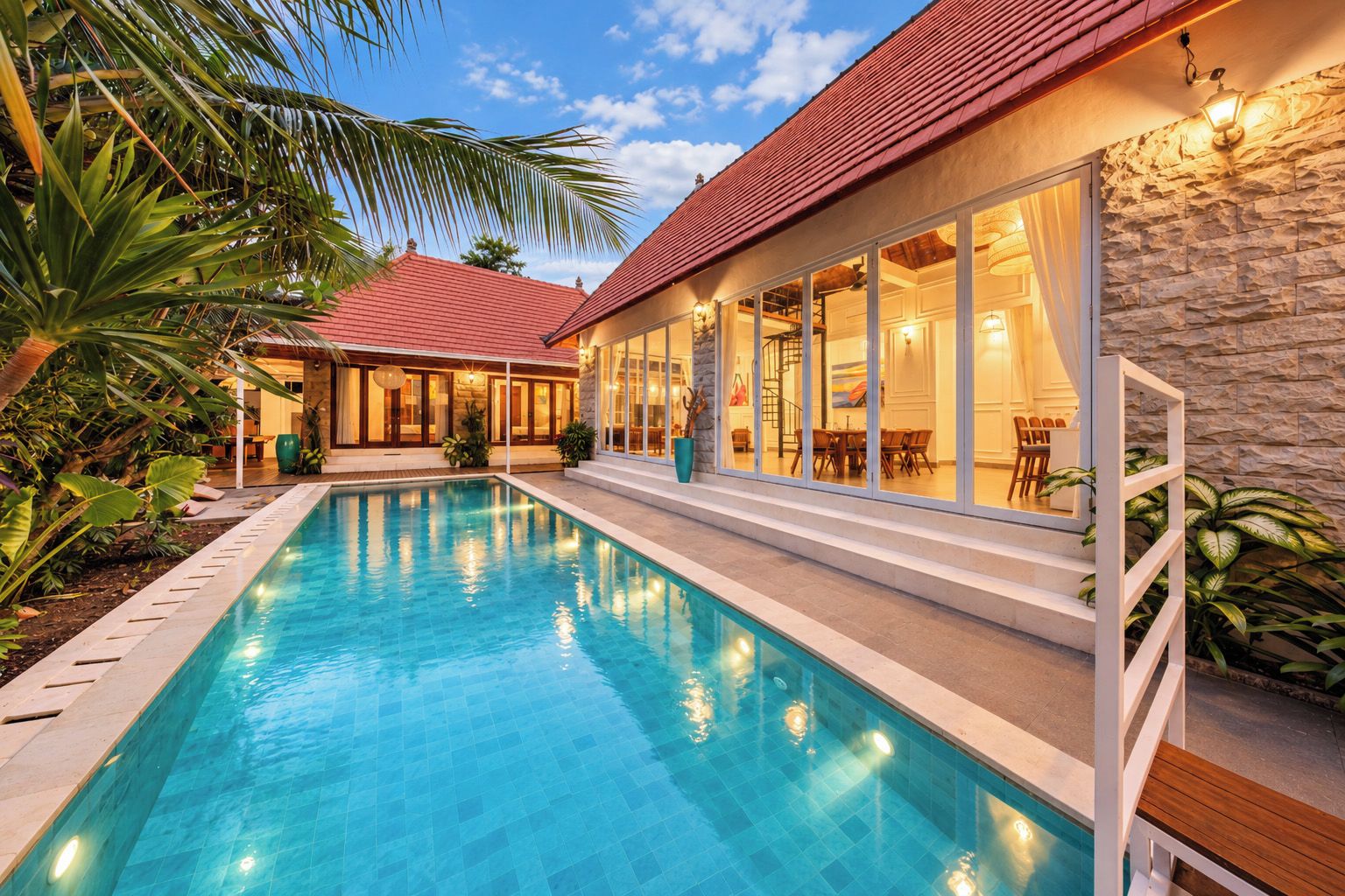 A swimming pool with a stone wall and a red roofed building with large windows.