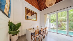 A modern kitchen with a large island, wooden chairs, a large mirror, and a view of a swimming pool. - Villa Naya Umalas (Kerobokan)