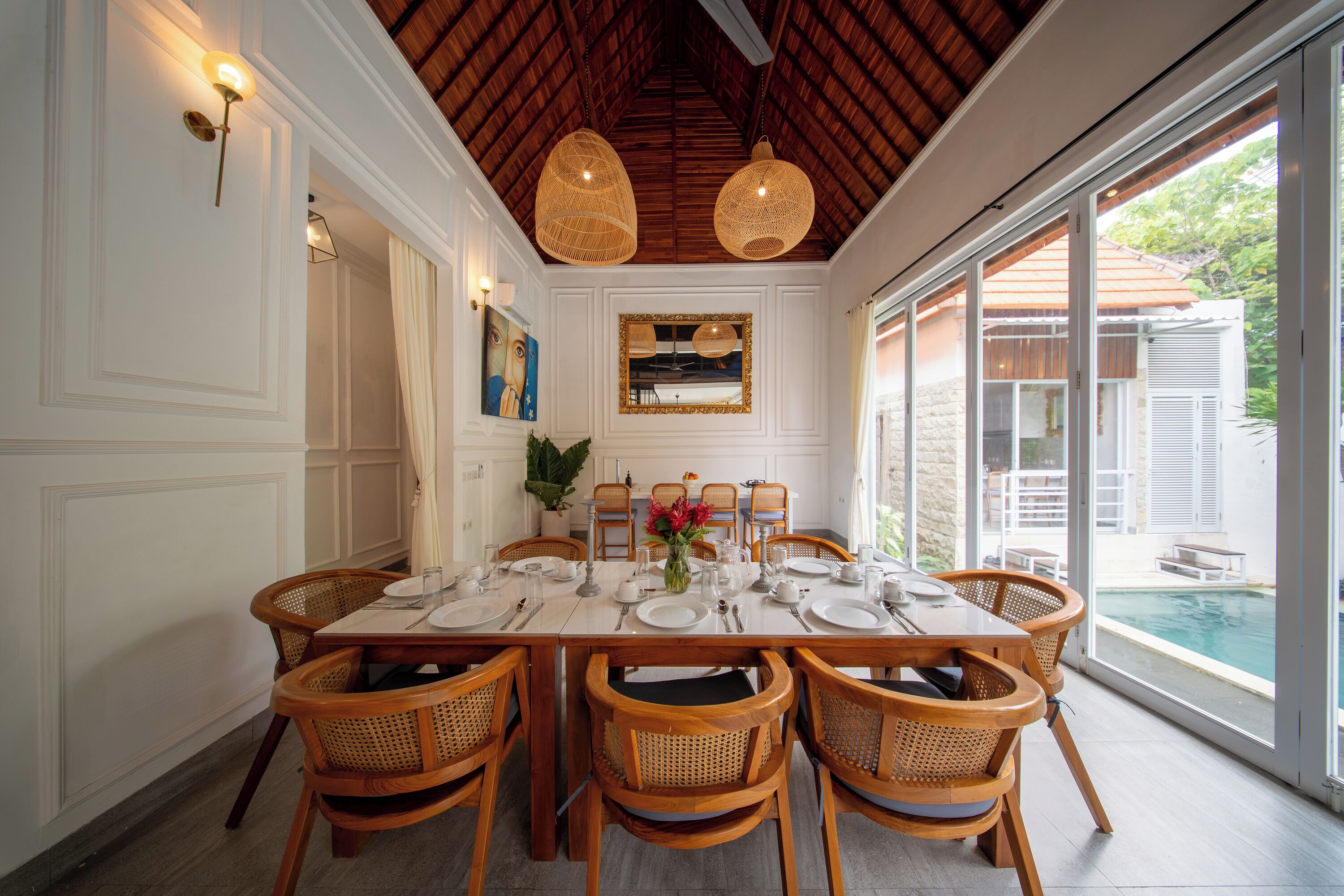 Un coin repas avec une table dressée pour six personnes, entouré de chaises en osier, d’un grand miroir et de luminaires suspendus.