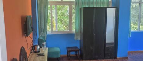 A room with a wooden floor, a ceiling fan, a large window with curtains, a wardrobe, a table with a kettle, and a bench.