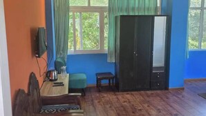 Una habitación con piso de madera, ventilador de techo, televisor montado en la pared, una mesa con hervidor, un banco y un armario.
