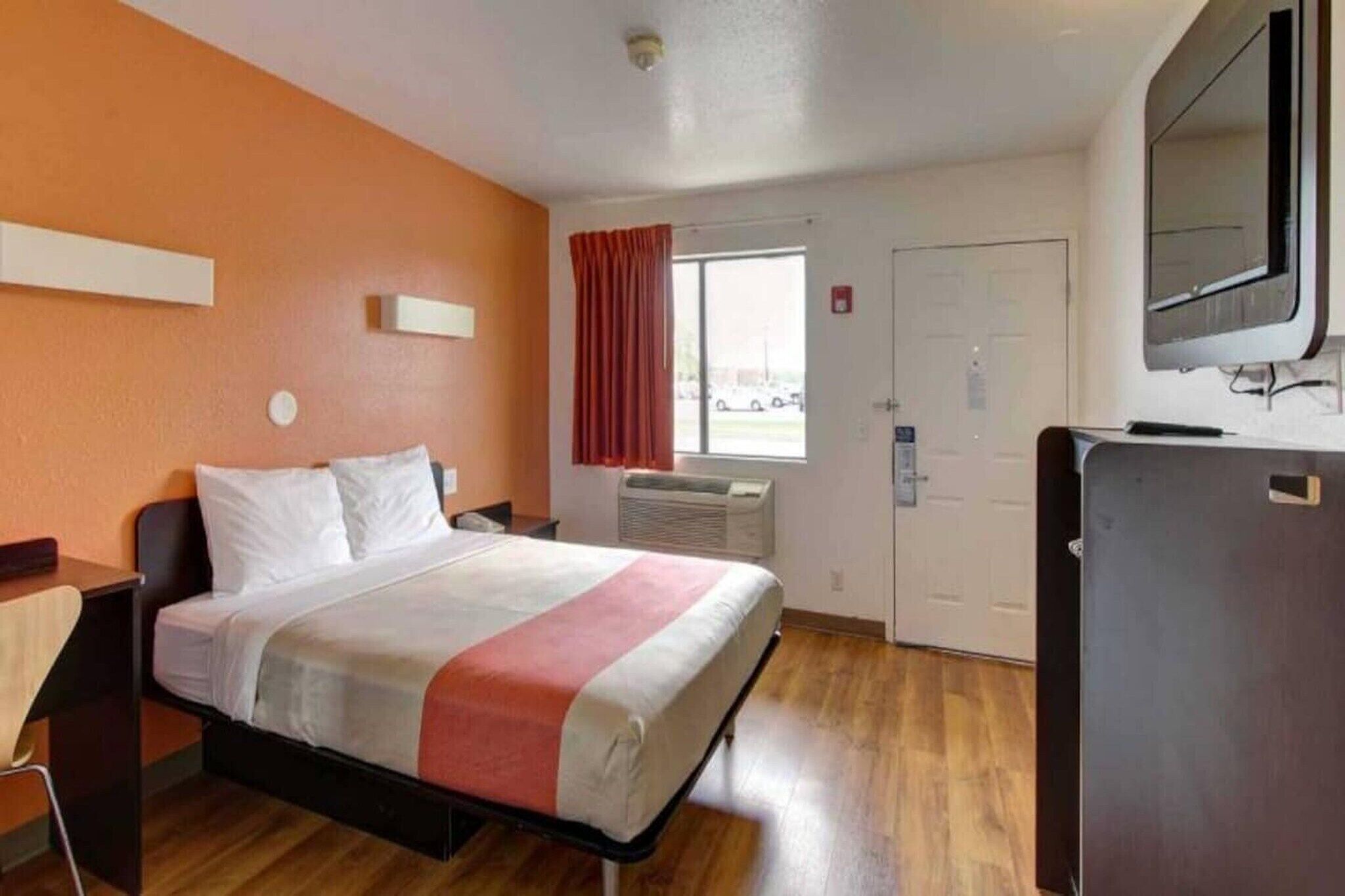 A single bed with a red and white bedspread, a desk with a chair, a window with curtains, a door, and a refrigerator.