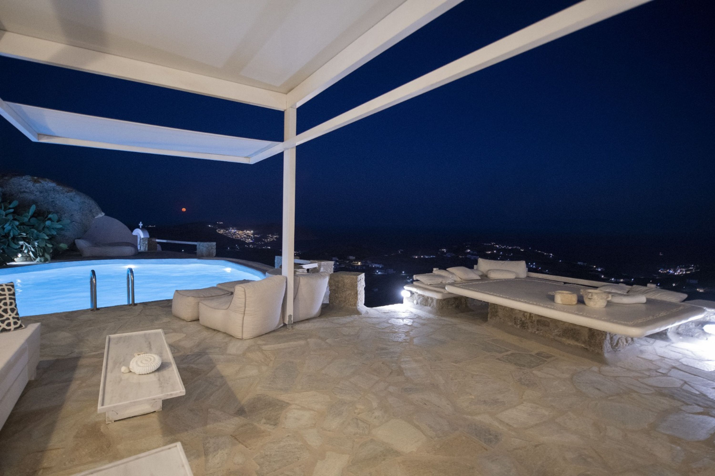Un espace piscine aménagé sur le toit, avec un sol en pierre, des meubles d’extérieur blancs et une vue sur la ville la nuit.